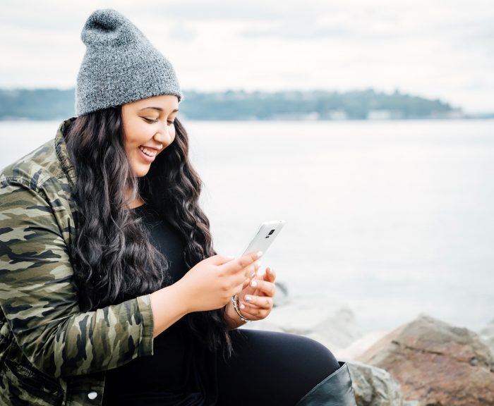 Woman smiling at her phone while resting on a rock by a lake.